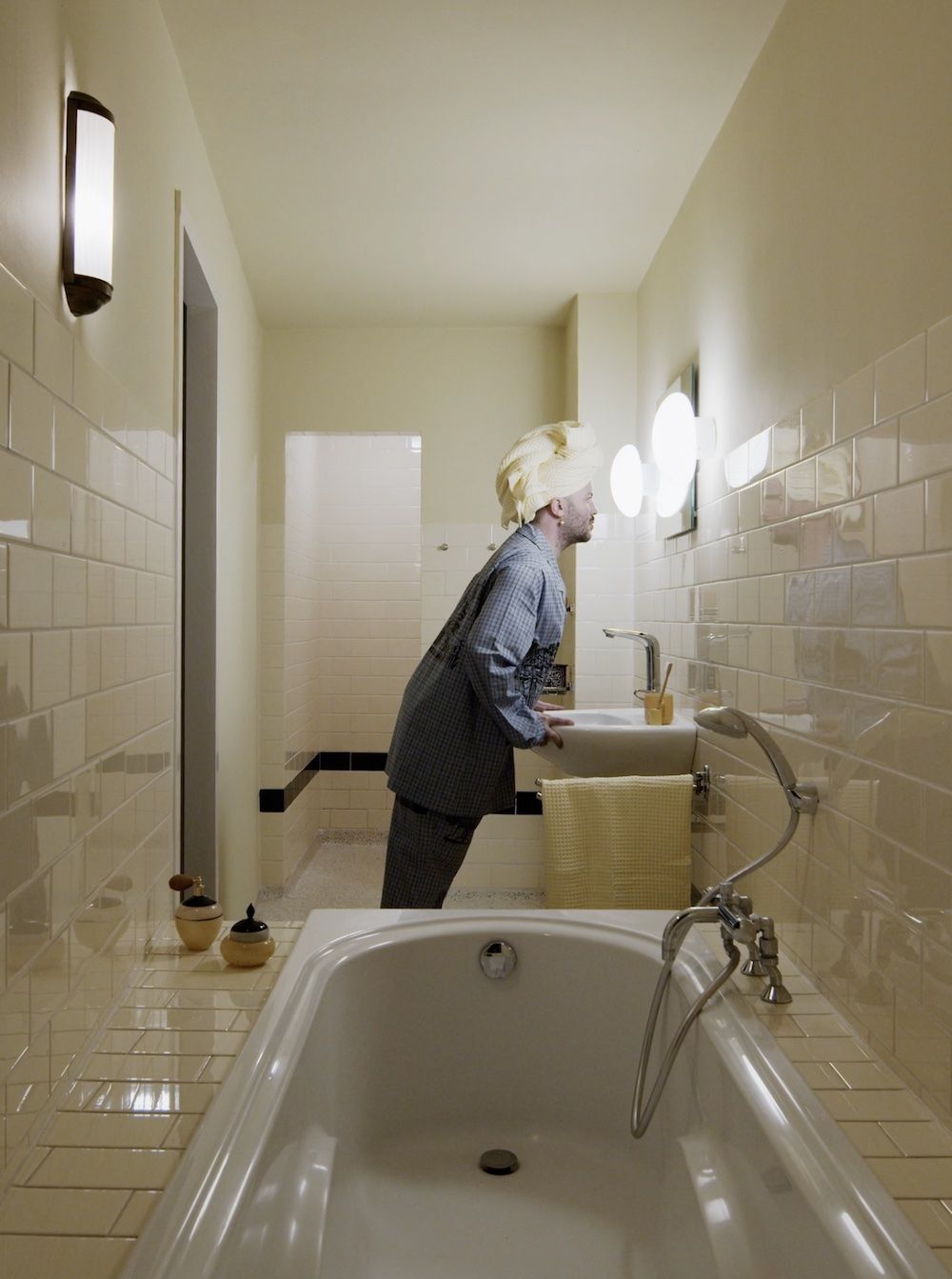 Een film-still uit Housewarming. Zijaanzicht van een man die inspecterend in de spiegel kijkt. Hij draagt een pyjama een handdoek op zijn hoofd.