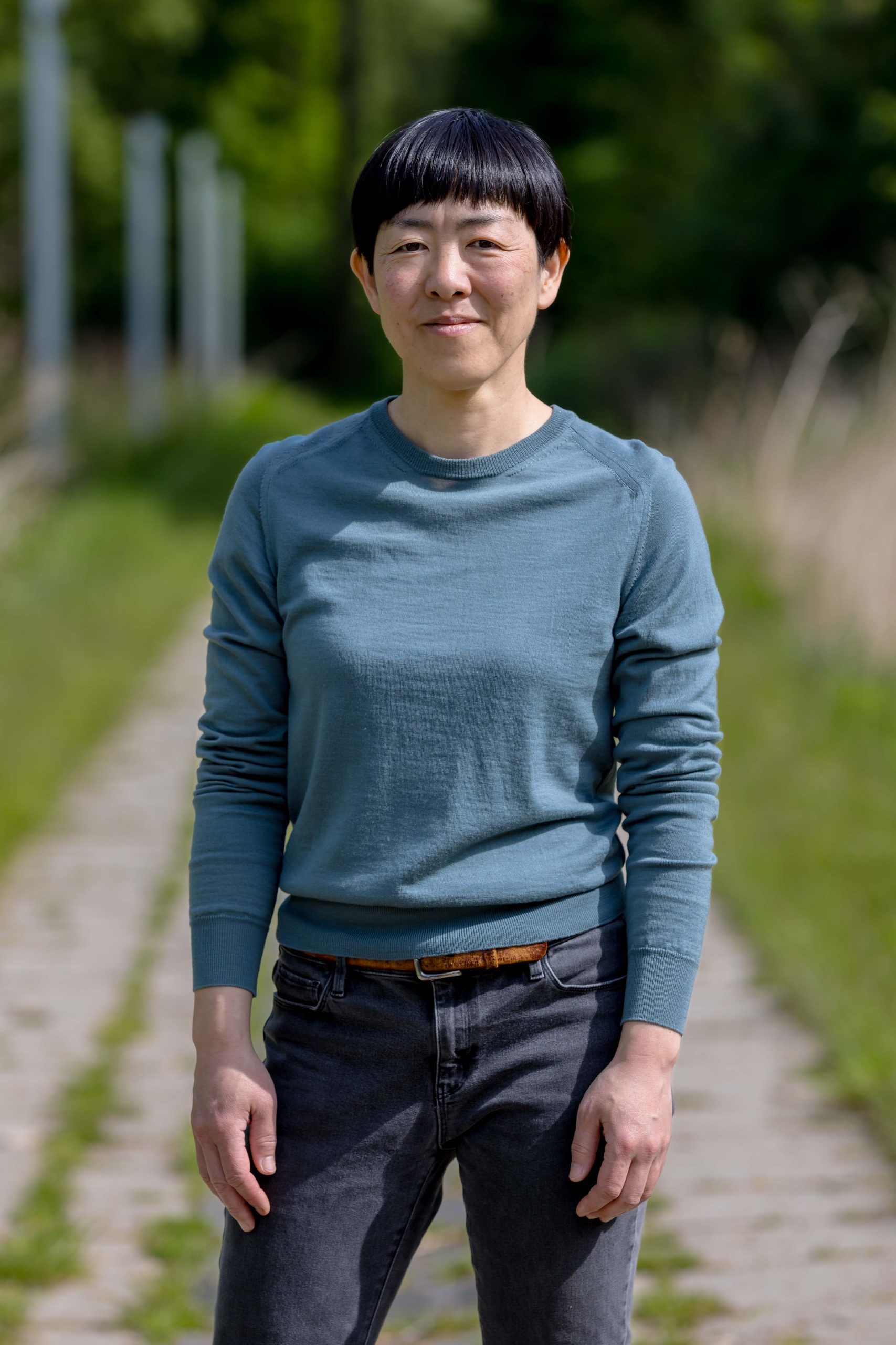 japanse vrouw met zwart haar en grauw blauw shirt en zwarte broek die recht in de camere kijkt. achtergrond van gras en beton
