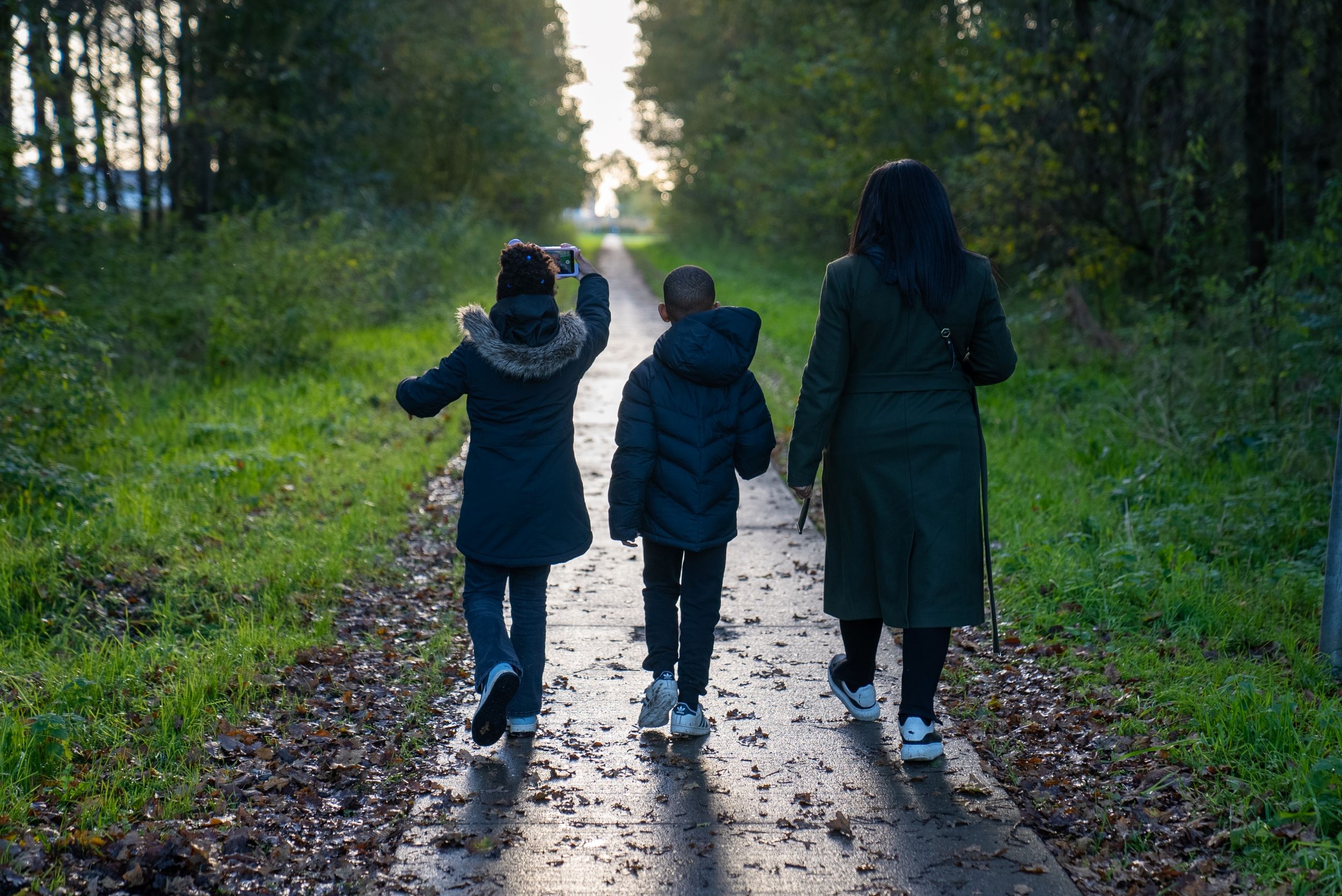 2 kinderen en hun moeder die over een asfaltweg lopen door een bos.