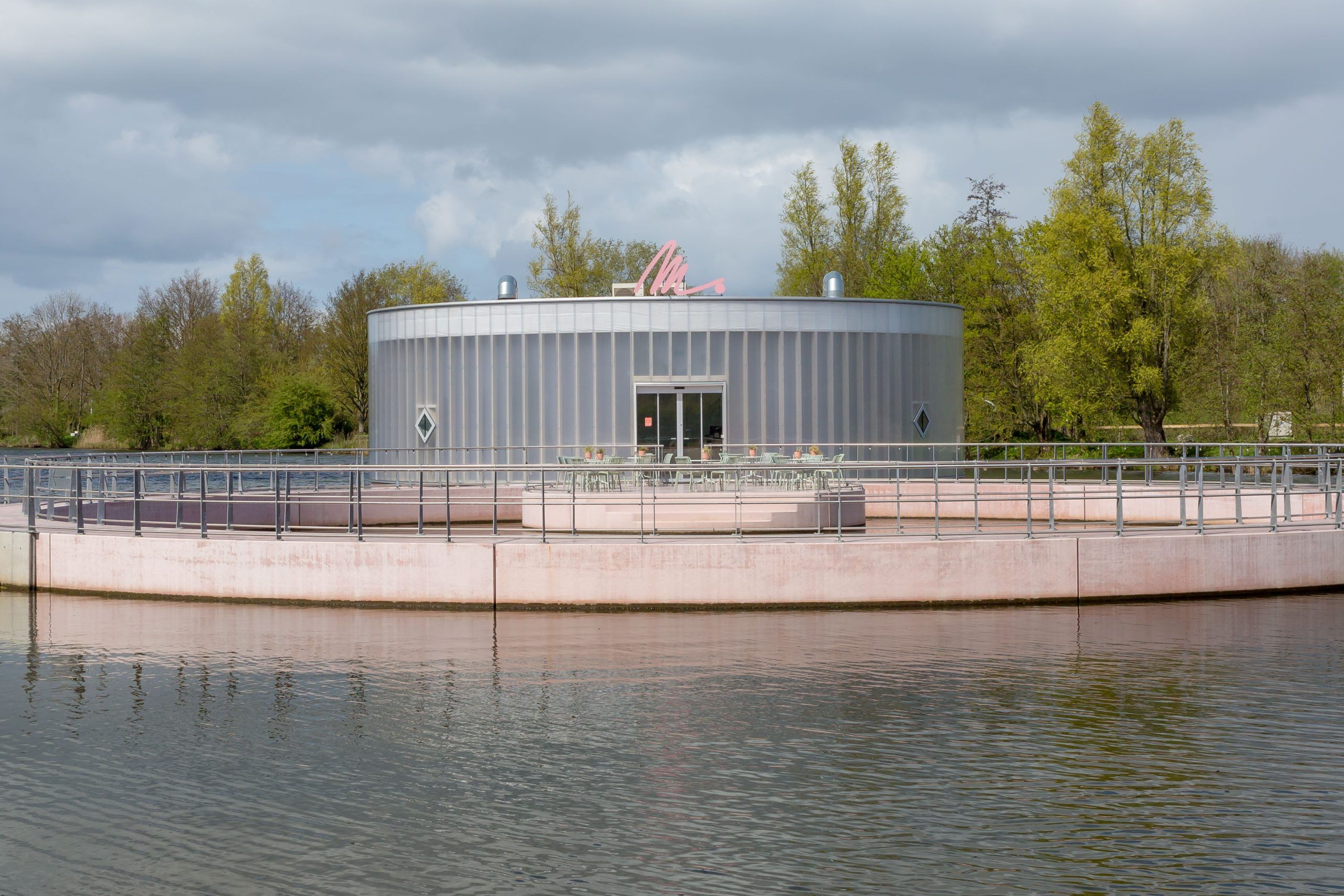 vooraanzicht van het kunstmuseum M. een rond paviljoen in het water met reling en een roze M. op het dak