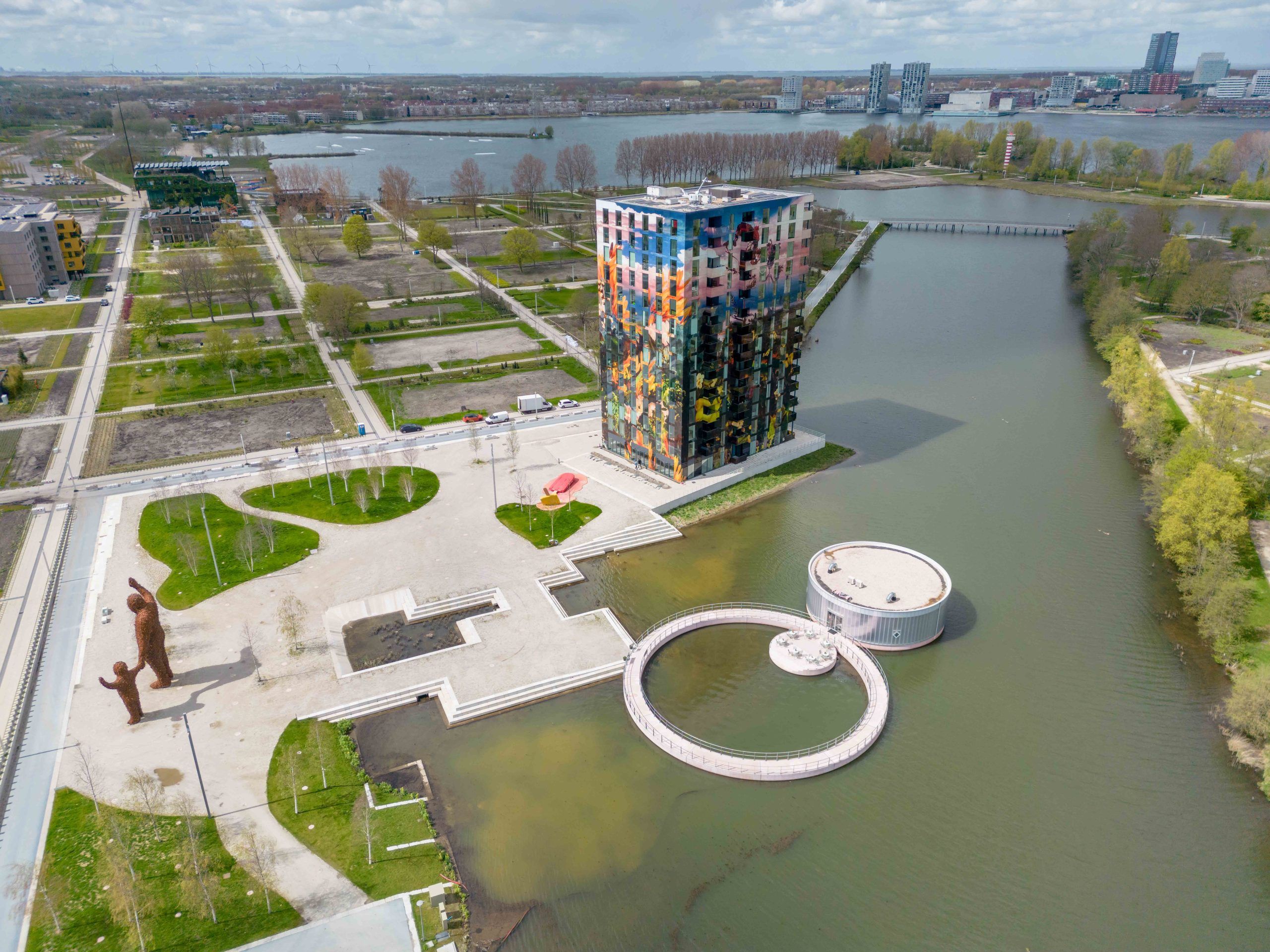 een drone foto vanuit hoog in de lucht die de skyline van Almere toont op de achtergrond, met daarvoor een grote kleurige woontoren en het ronde paviljoen van M. in het water.
