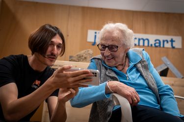 witte man met snor en lang sluik haar laat oude witte mevrouw in rolstoel een filmpje zien op een telefoon