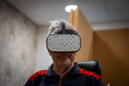 oude witte man met grijs haar en een VR bril op zijn ogen die de camera inkijkt