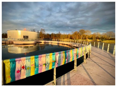 veel verschillende kleuren zijden linten aan een reling voor een rond museumgebouw. op de linten zijn door kinderen hun wensen voor dit jaar geborduurd