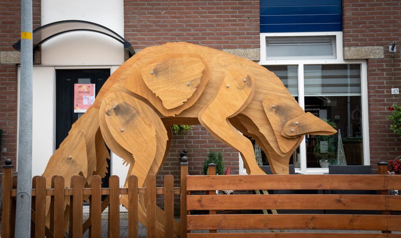 Een houten vos wat met zijn achterpoten in een voortuin staat en met zijn voorpoten in de voortuin van de buren.