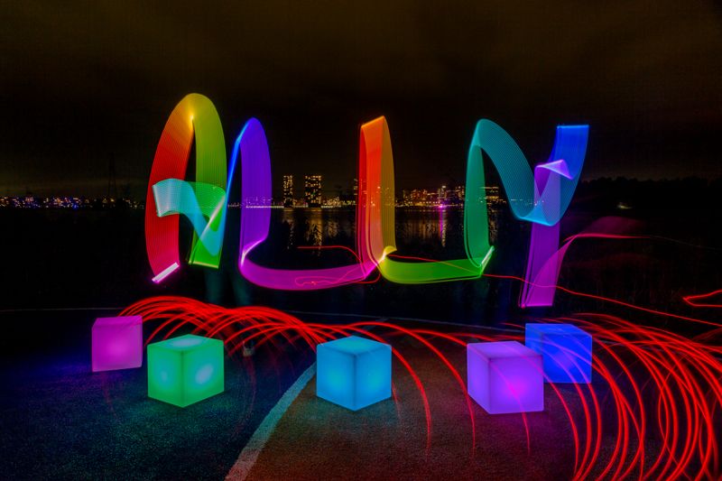 lichtkunstfoto met heel veel verschillende kleuren. er is ALLY geschreven met hoofdletters in diverse kleuren. onder de letters staan 5 gekleurde kubussen die licht geven. ieder in een andere kleur, van paars naar groen naar blauw naar paars naar donker blauw