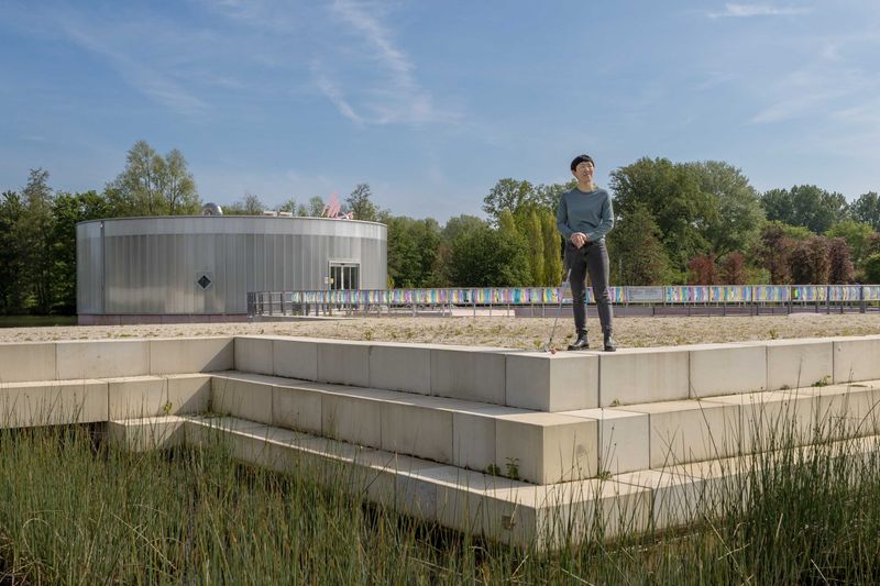 japanse vrouw met zwart haar en grauw blauw shirt en zwarte broek staat op een betonnen trap, kijkt naar rechts. op de achtergrond het kunstmuseum M. een rond gebouw met een M. op het dak. verder veel gras in de voorgrond en beton in de achtergrond. helemaal achterin het beeld zijn bomen